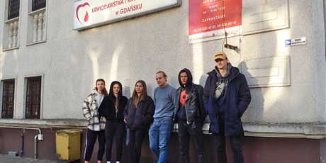 Powiększ grafikę: 6 wychowanków stojących przy budynku RCKiK. Nad młodzieżą znajduję się tablica z napisem „Regionalne Centrum Krwiodawstwa i Krwiolecznictwa w Gdańsku” oraz tablica z godzinami przyjmowania dawców