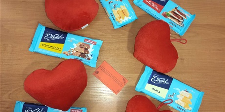 Powiększ grafikę: Na zdjęciu widać pięć czekolad, cztery pluszowe serduszka w kolorze czerwonym i karteczka z życzeniami. Wszystko leży na stoliku
