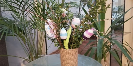 Powiększ grafikę: Na pierwszym planie dekoracja  w wazonie bukszpan oraz dekoracyjne jajka pisanki, za dekoracją kwiaty doniczkowe.