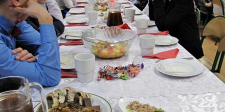 Powiększ grafikę: Na pierwszym planie uroczyście ozdobiony stół z wigilijnymi potrawami, dookoła siedzą wychowankowie. W tle grupa wychowanków dająca występ artystyczny.