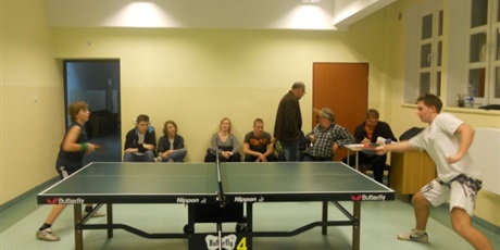 Powiększ grafikę: Sala  do pin-ponga, w której  toczy się zażarty pojedynek zawodników, któremu przygląda się z ławki widownia.